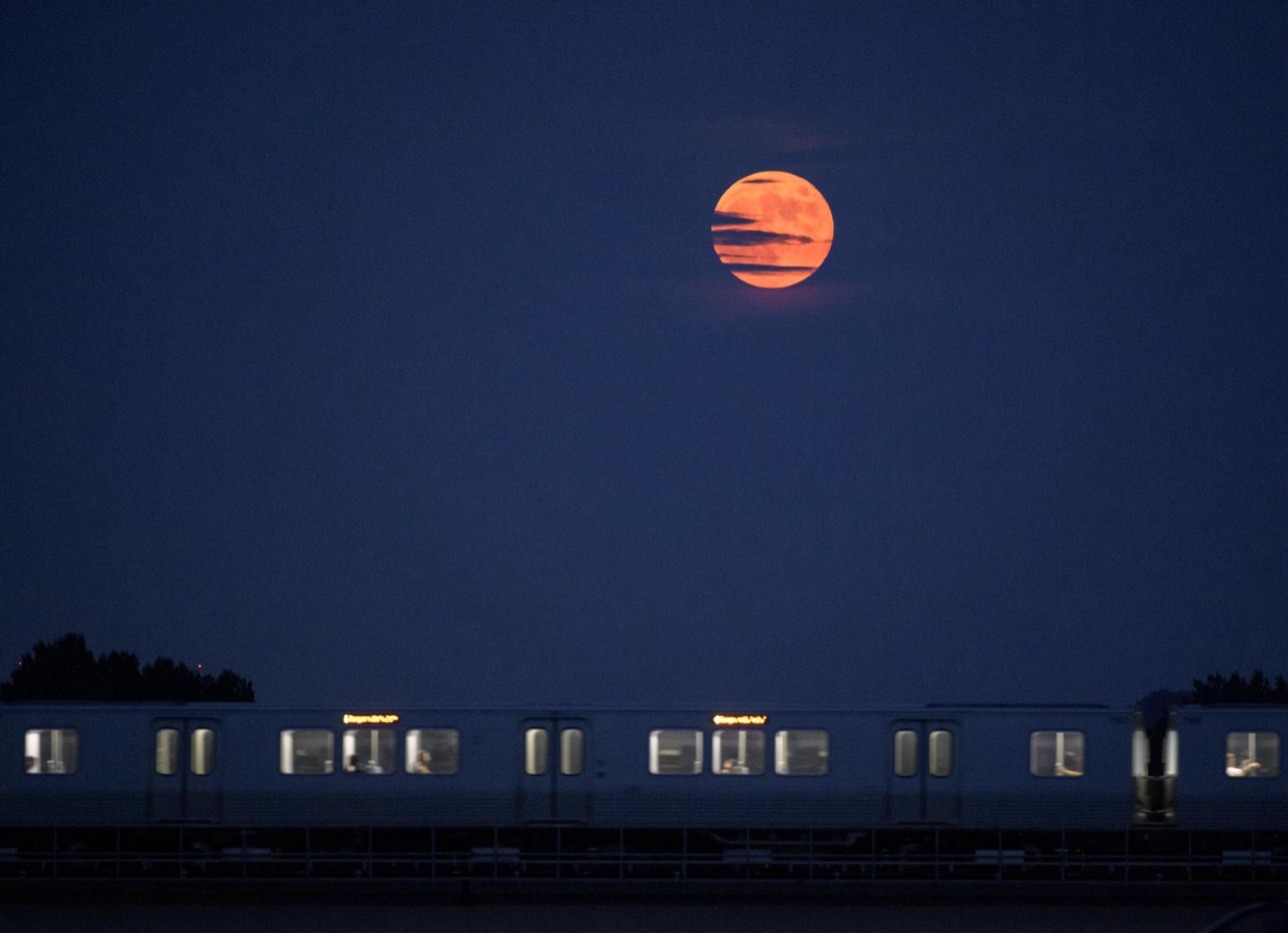A large full Moon rising over a river while a lit commuter train crosses a bridge in the foreground