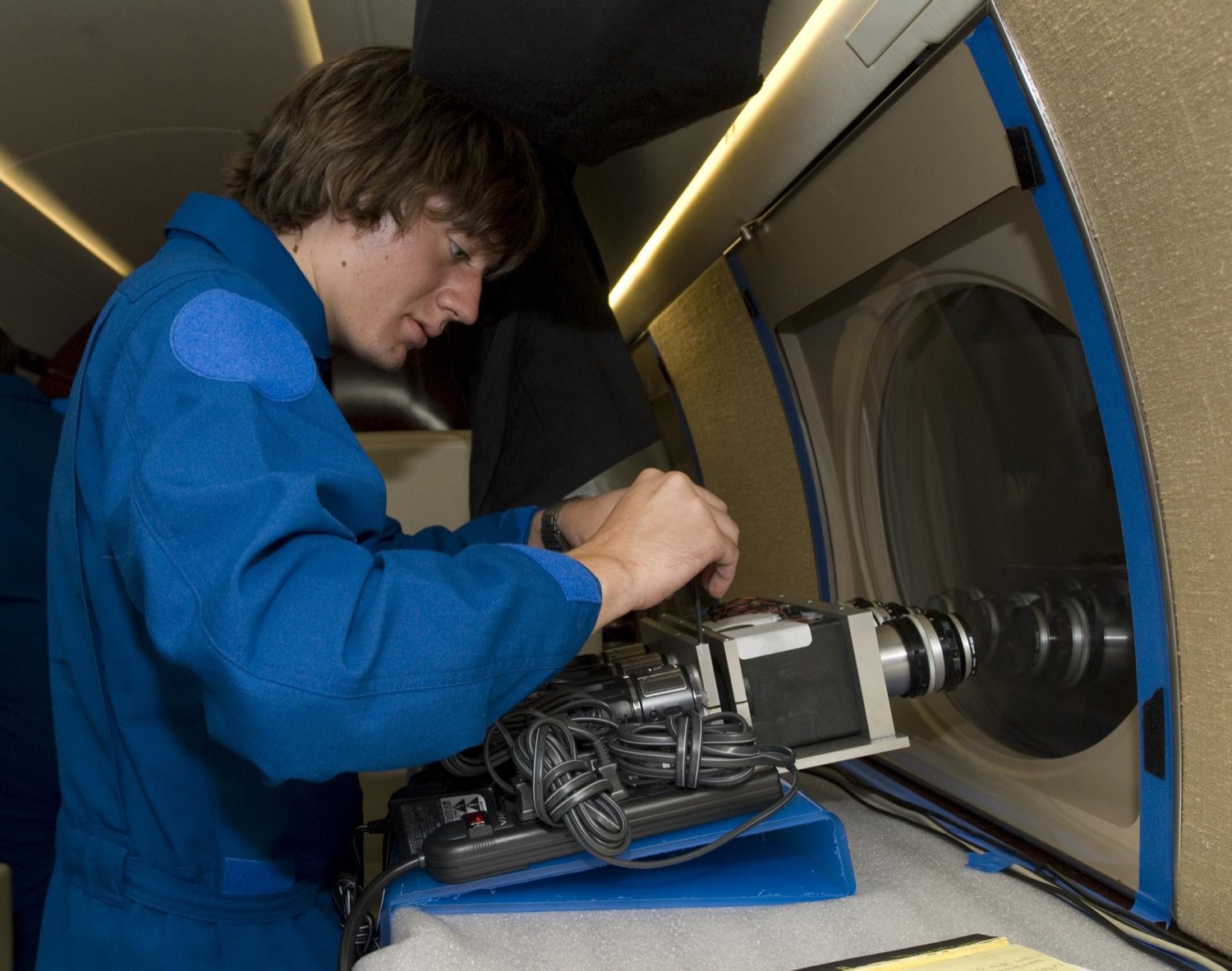 A young researcher in a blue flight suit adjusts a camera pointed out of an aircraft window